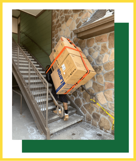 A person carrying a large, strapped-together box up a concrete outdoor staircase next to a stone wall.