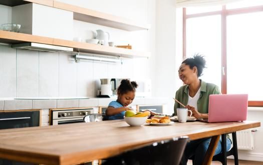 A person with a pink laptop and a child sit at a kitchen table, smiling while eating snacks.