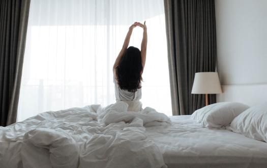 A person sitting on a bed in a bright bedroom, stretching their arms toward a large window.