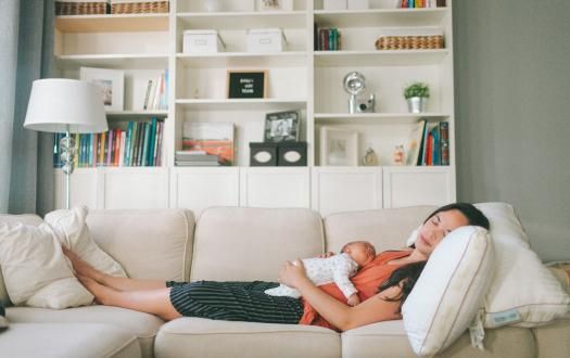 A person rests on a sofa while holding a sleeping infant on their chest, with a bookshelf in the background.