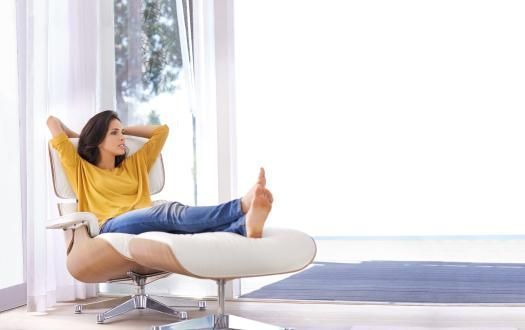 A person wearing a yellow shirt and jeans relaxing in a white lounge chair with an ottoman in a bright, sunlit room.