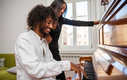 A person plays a wooden piano while another person stands beside them, smiling.