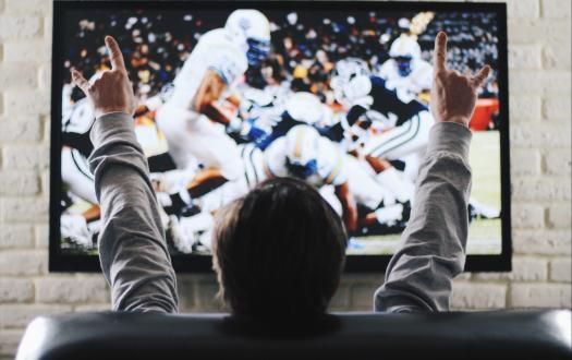 A person sits in a chair facing a TV showing a football game, arms raised with hands making a rock-and-roll gesture.