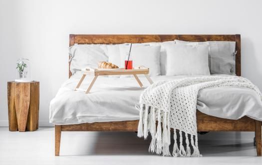 A rustic wooden bed with white bedding, a chunky knit throw, a small breakfast tray, and a matching wooden nightstand.