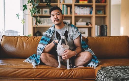 A person sitting cross-legged on a leather couch, draped in a blue patterned blanket, holding a small dog.