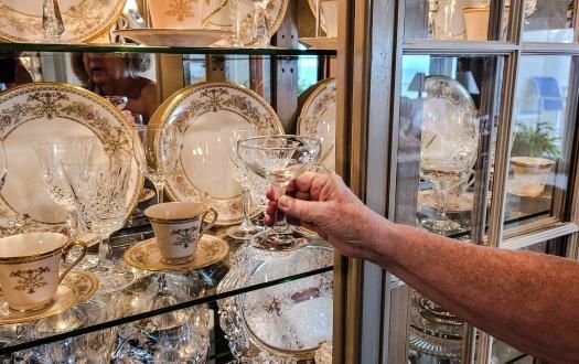 A person's hand reaches into a glass curio cabinet filled with fine china plates and crystal glassware.