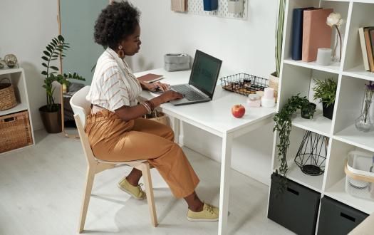 A person sits at a white desk in a bright, organized home office, typing on a laptop with an apple nearby.