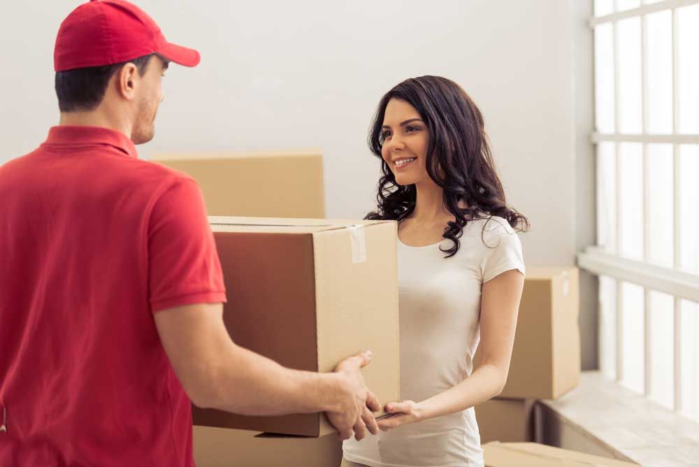 A courier in a red uniform handing a cardboard delivery box to a smiling person in a bright, sunlit room with boxes.