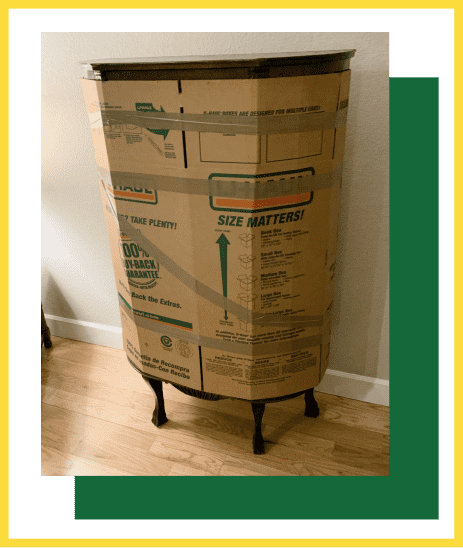 A vintage wooden side table with cabriole legs is wrapped in flattened cardboard boxes and secured with packing tape.