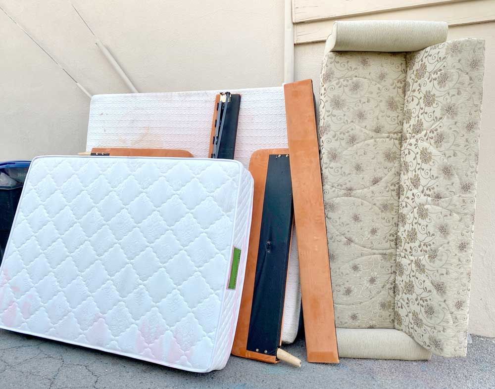 Discarded furniture, including a mattress, a patterned upholstered headboard, and wooden frame parts, leaning on a wall.