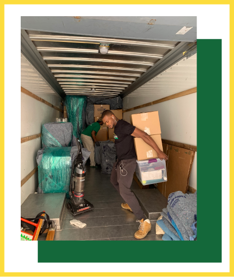 A person wearing a black shirt and tan boots carries stacked cardboard boxes and a plastic bin inside a moving truck.