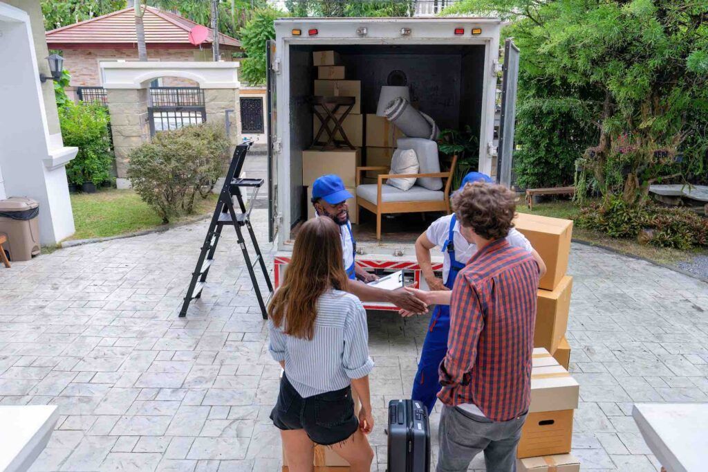 Two people shake hands with a moving professional in front of a truck loaded with furniture at a residential property.