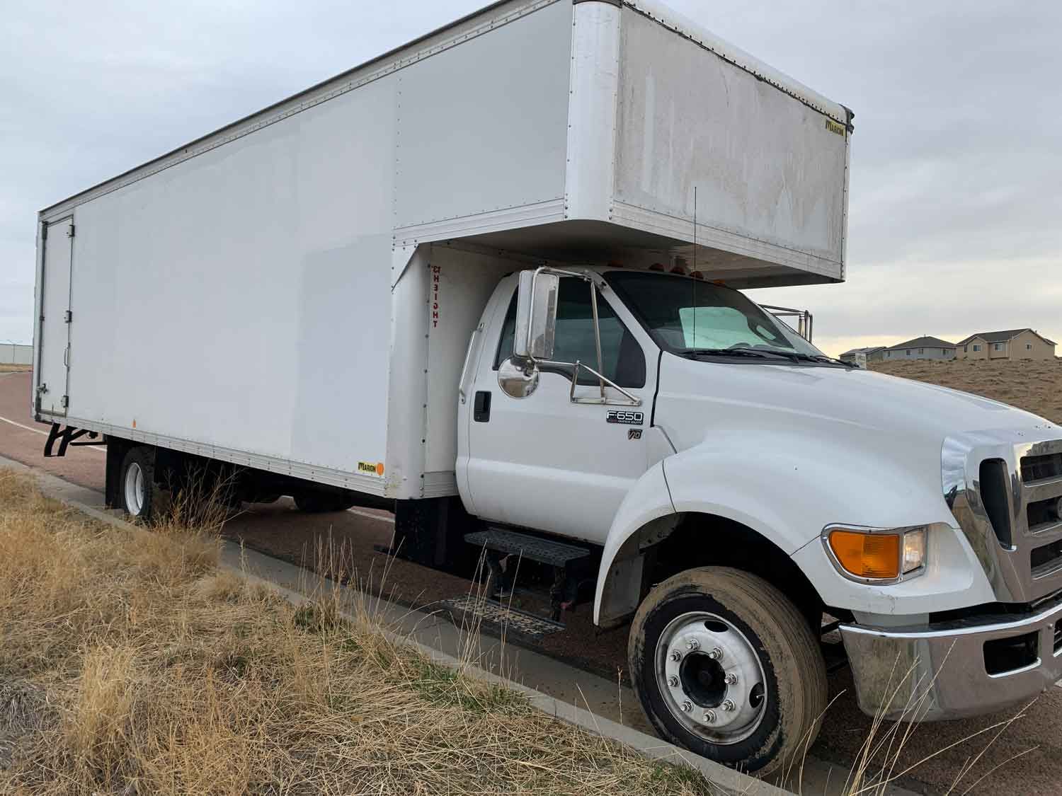 A white box truck parked on a roadside with dry grass under a cloudy sky.