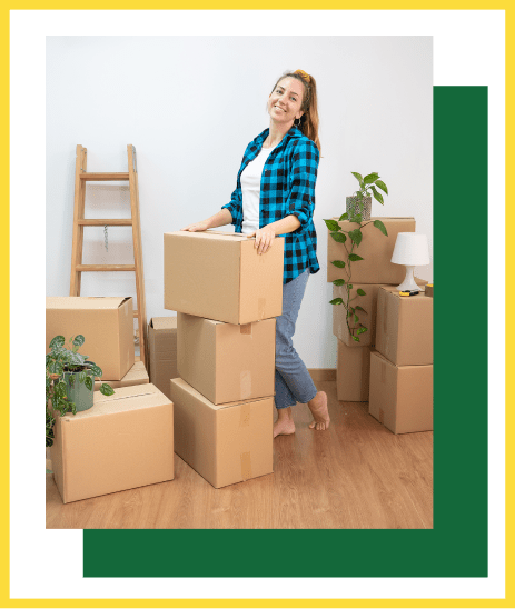 A person in a blue plaid shirt stands smiling in a room, leaning against a stack of cardboard moving boxes near a ladder.