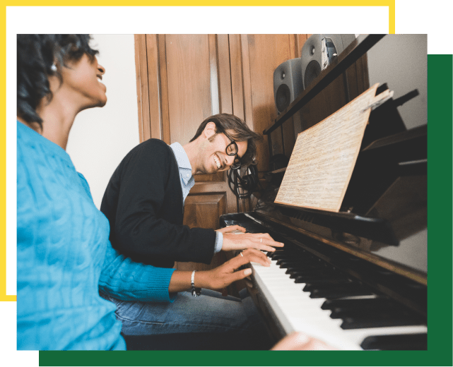 A person in a blue sweater and another in a black cardigan play a piano together, both smiling.