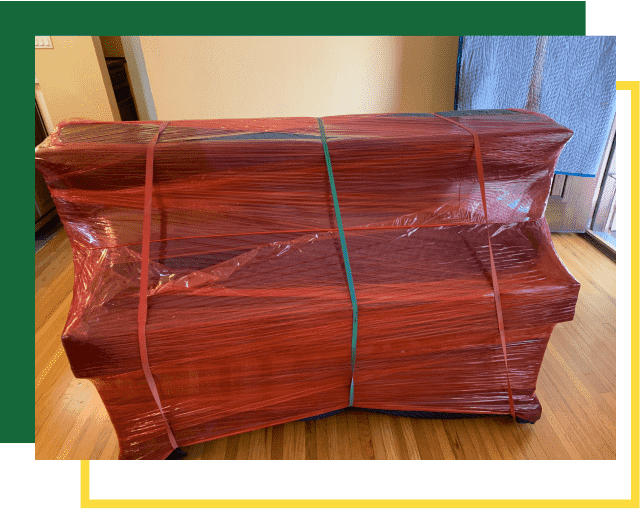 An upright piano wrapped in protective red plastic film and secured with straps, standing on a hardwood floor.