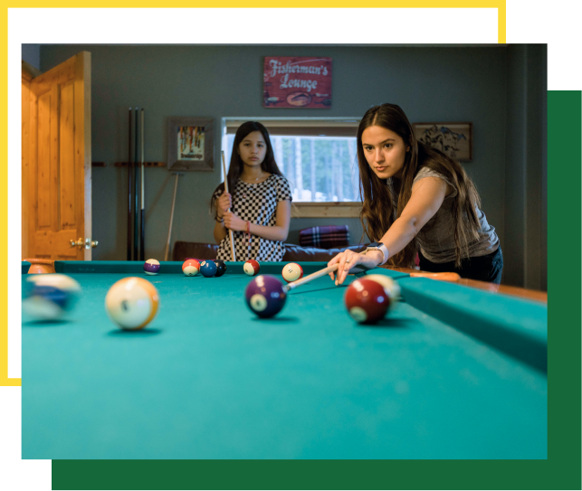 Two young people play a game of pool on a green table; one is aiming a cue stick while the other watches in the background.