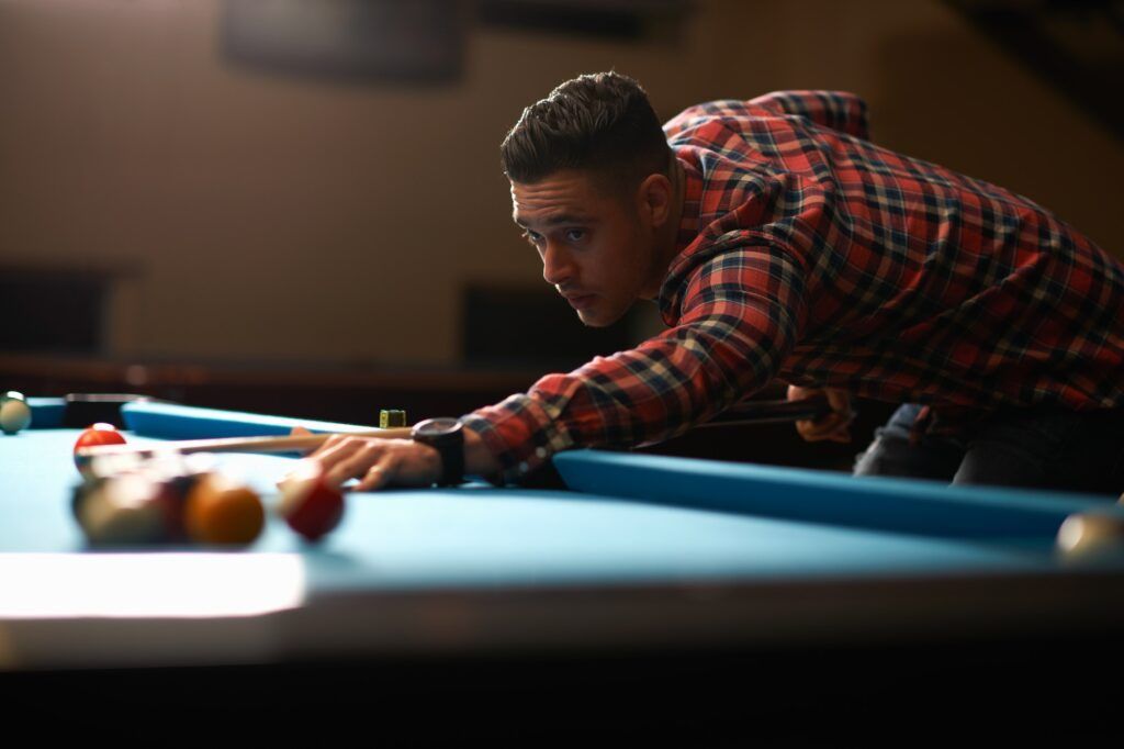 A person in a red plaid shirt leans over a blue pool table, aiming their cue stick at a cluster of colorful billiard balls.