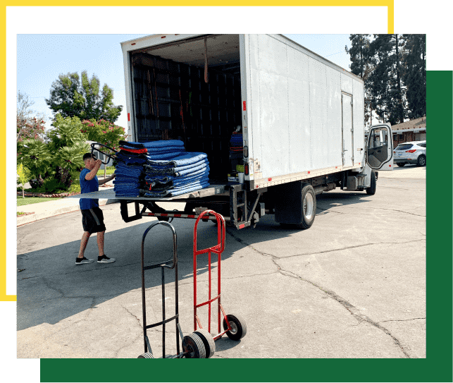 A person loads blue furniture moving pads onto a large white moving truck parked on an asphalt street.