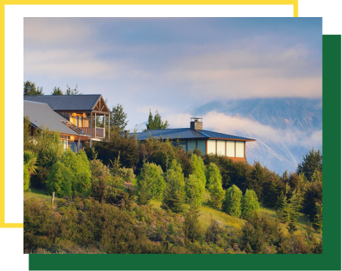 Two houses situated on a grassy, tree-covered hillside with a large mountain in the background under a cloudy sky.