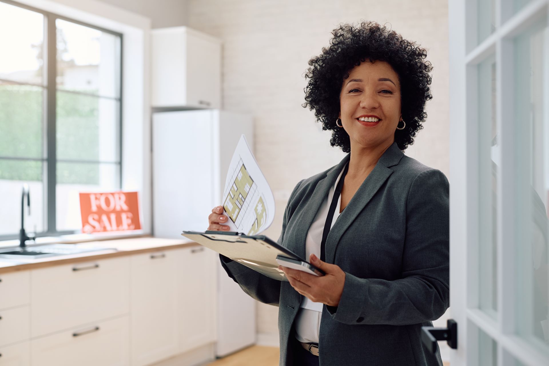 A woman in a suit is holding a clipboard in front of a for sale sign.