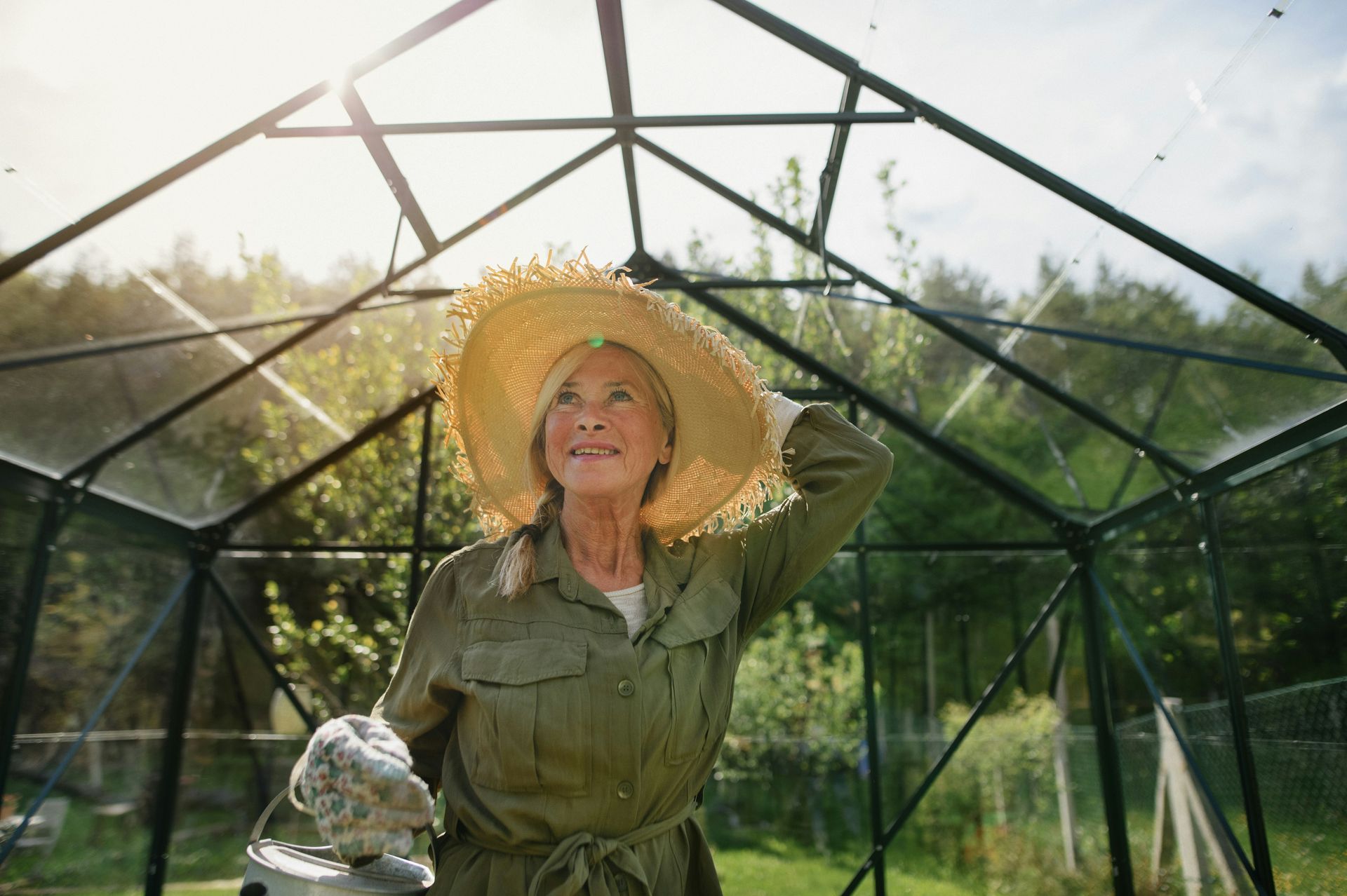 An elderly woman wearing a straw hat is standing in a greenhouse.