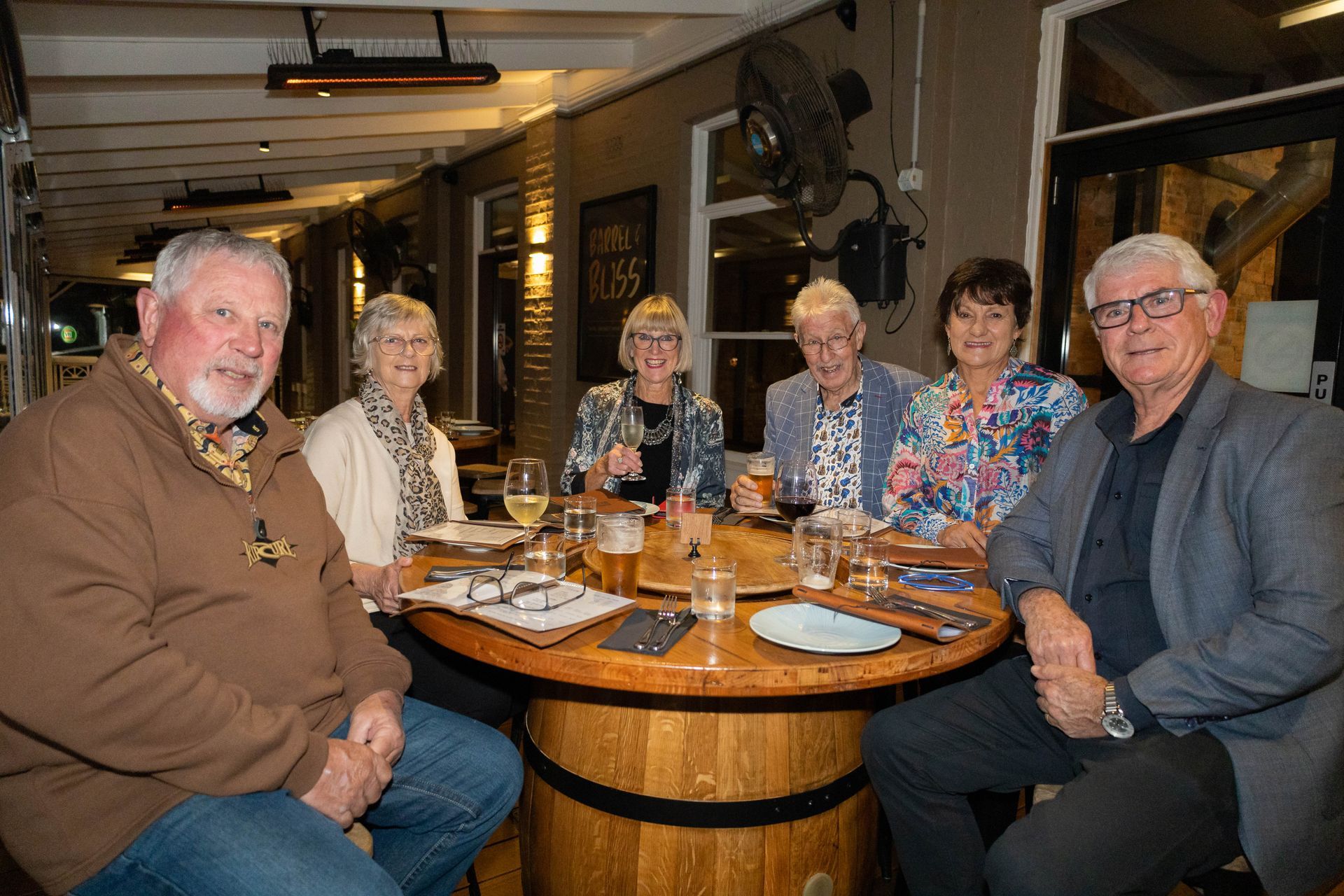 A group of six people sitting around a round wooden table at a restaurant, enjoying drinks and food.