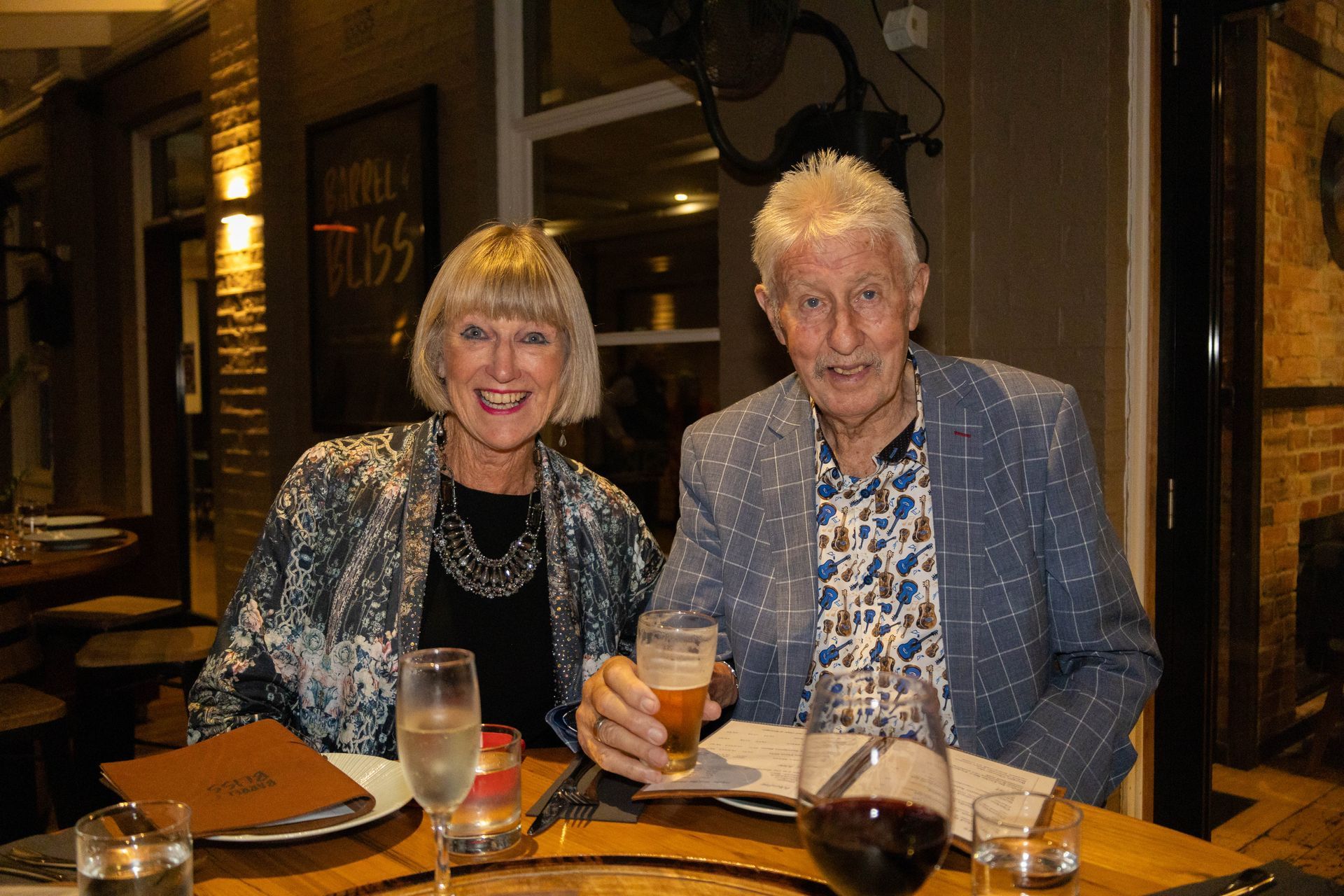 A smiling older couple at a restaurant table, the man holding a beer, the woman with a champagne flute.