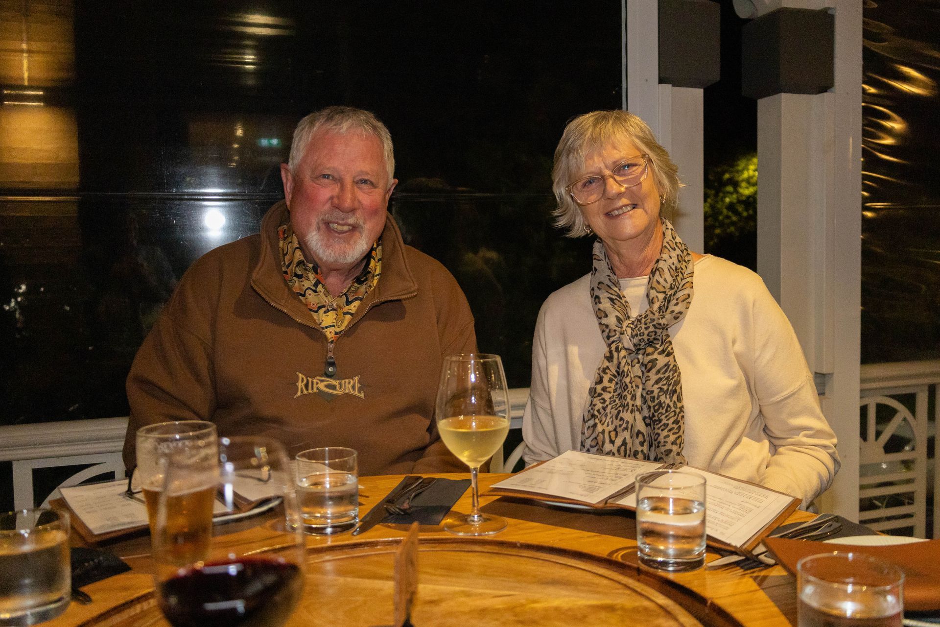 Two people, smiling, at a table with drinks and menus. Nighttime outdoor setting.