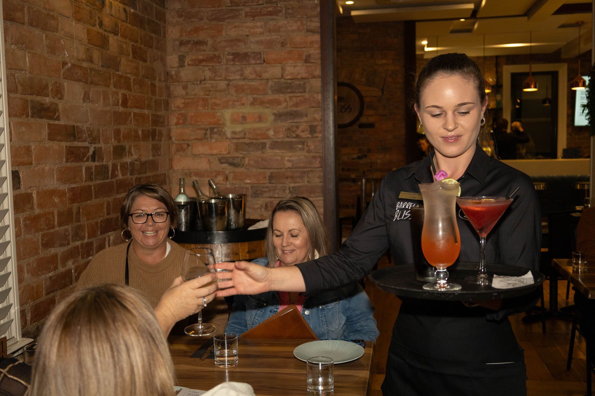 A waitress serves cocktails to a group of women at a restaurant. Brick wall background.