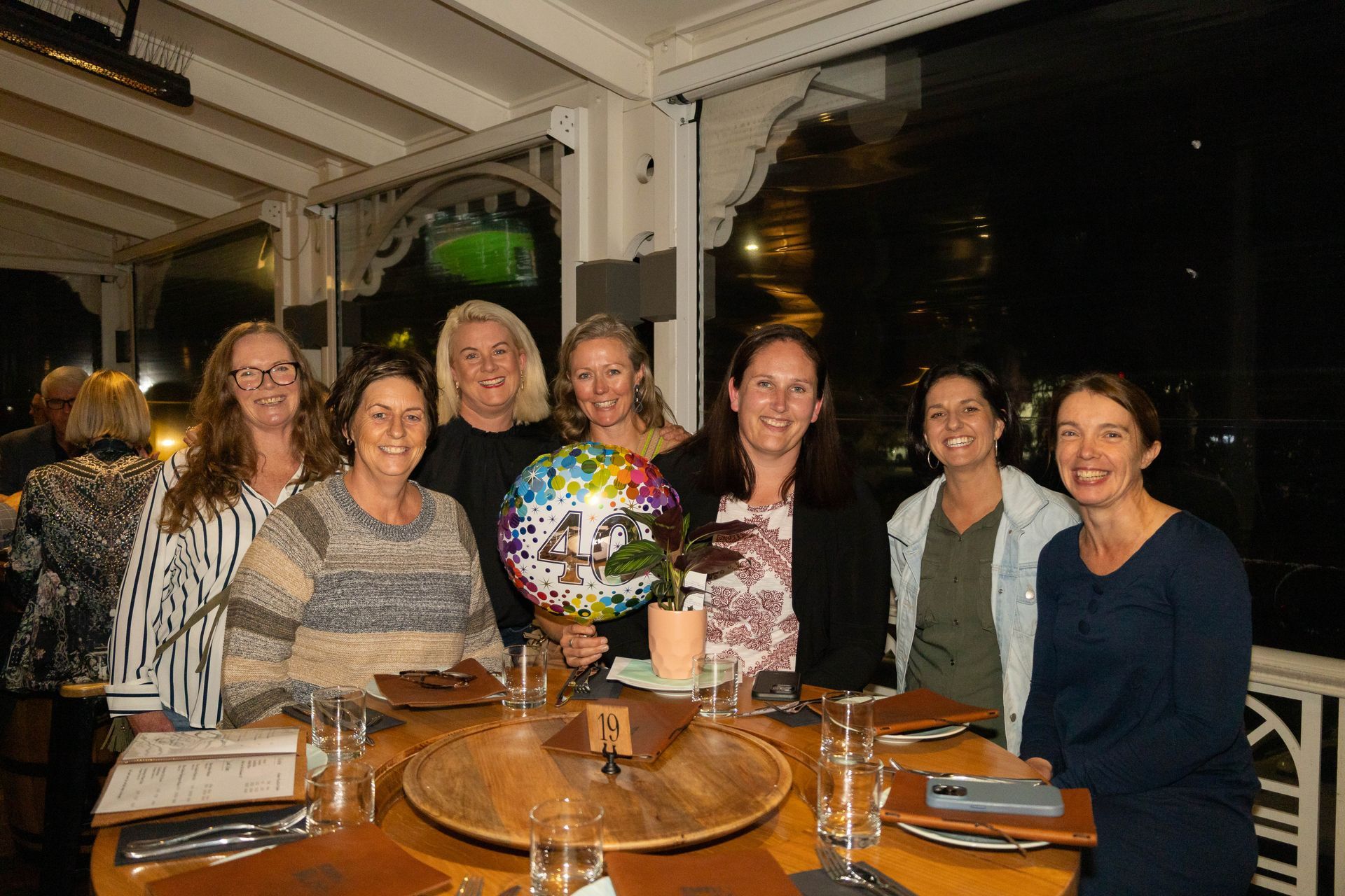 Group of women at a restaurant table, some holding a balloon; nighttime setting.