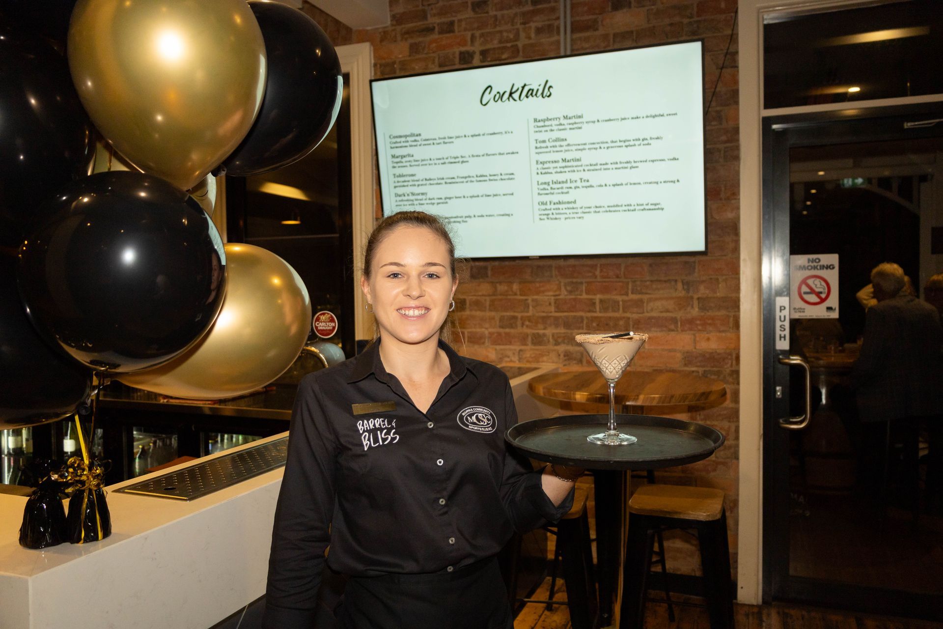 Bartender holding cocktail on a tray at bar with black and gold balloons and cocktail menu.