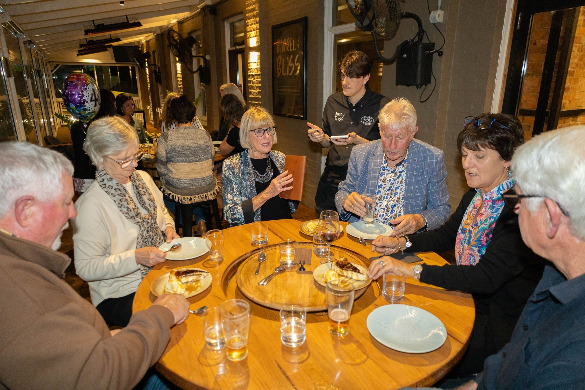 A group of senior adults seated around a wooden table at a restaurant, eating and socializing.