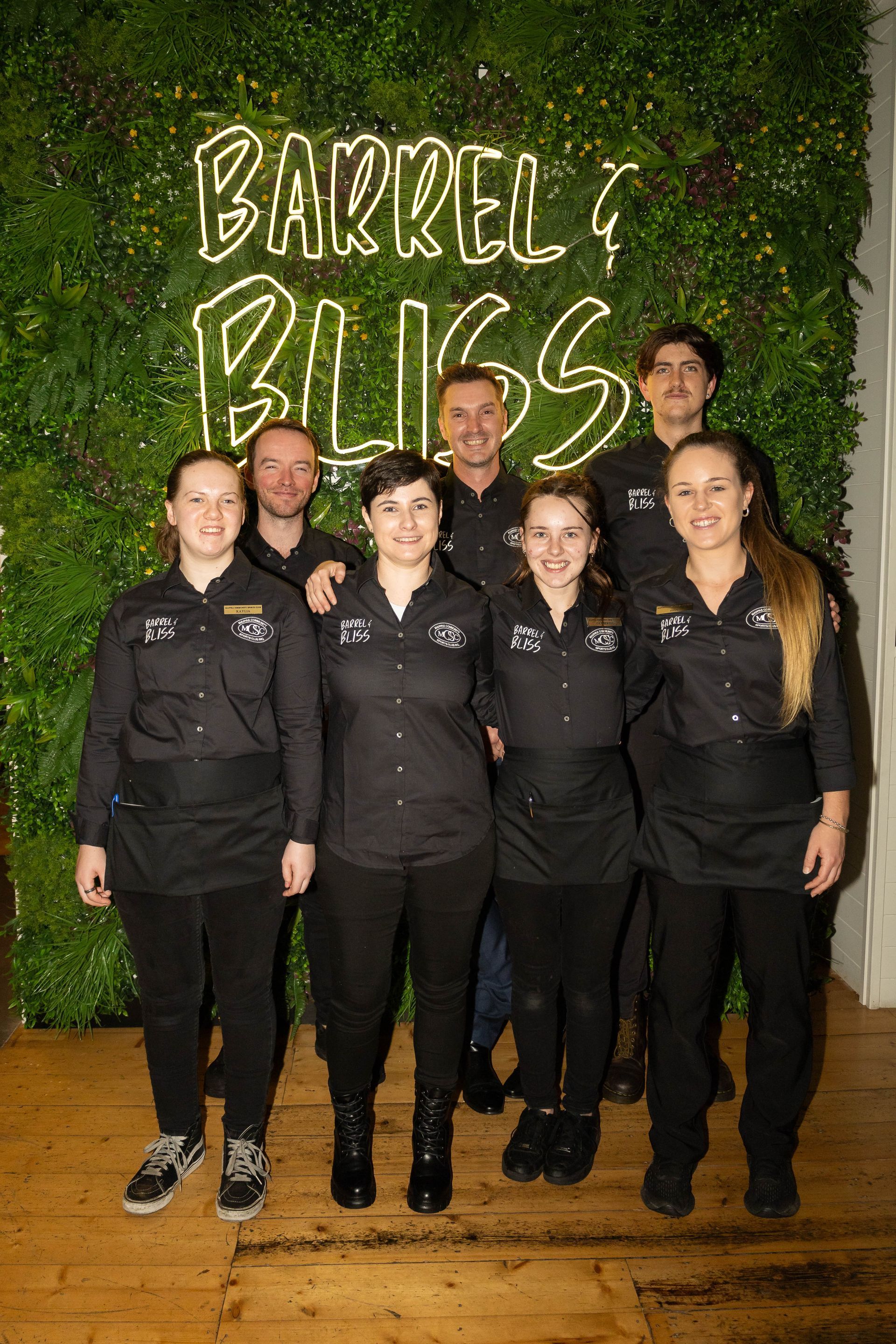 Group of people in black uniforms stand in front of a green wall with a neon sign that says 