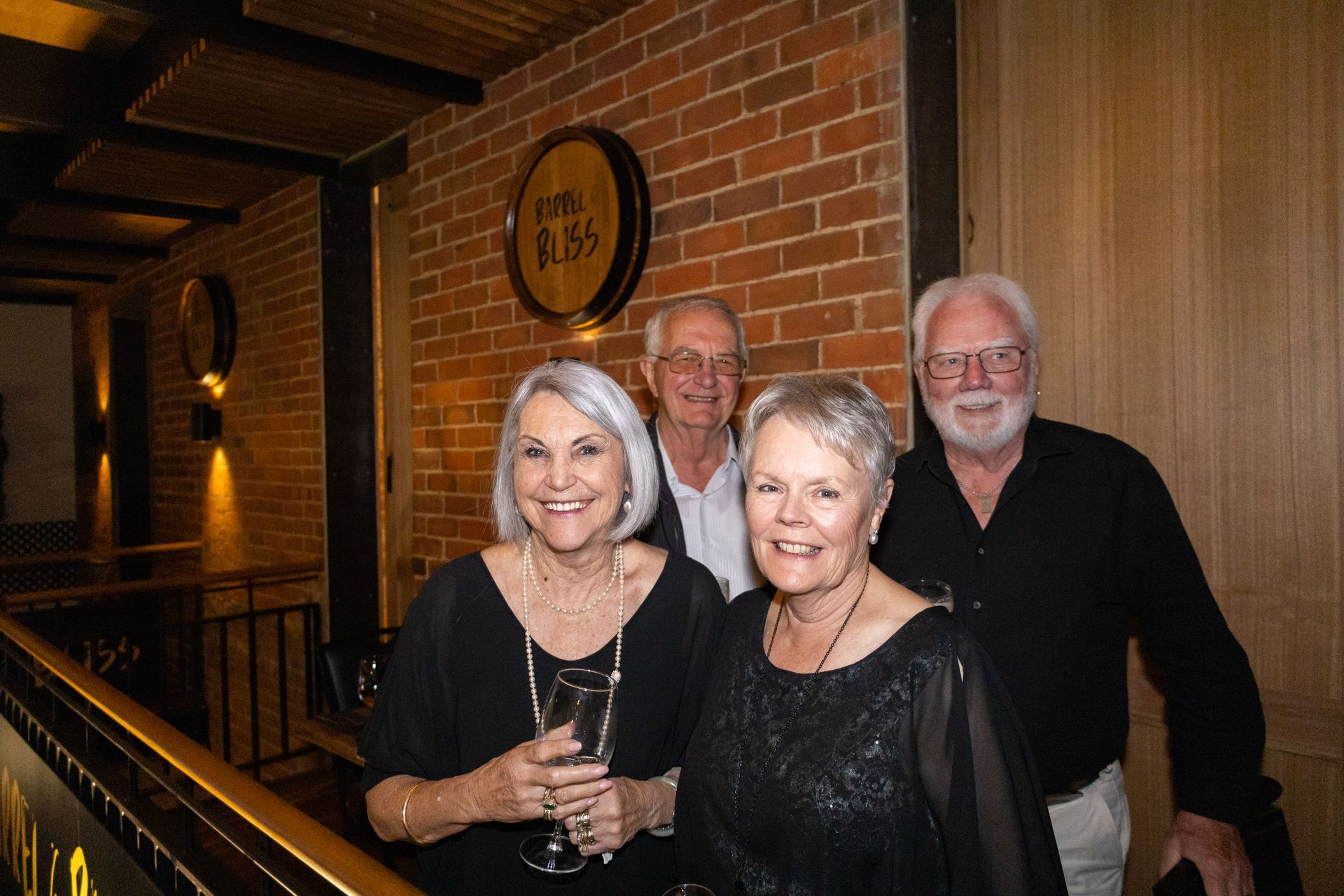 Four people smiling at a camera, standing in front of a brick wall with a barrel sign.