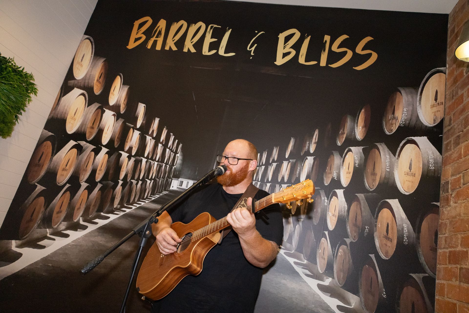 Man playing acoustic guitar at Barrel & Bliss, backed by mural of barrels.