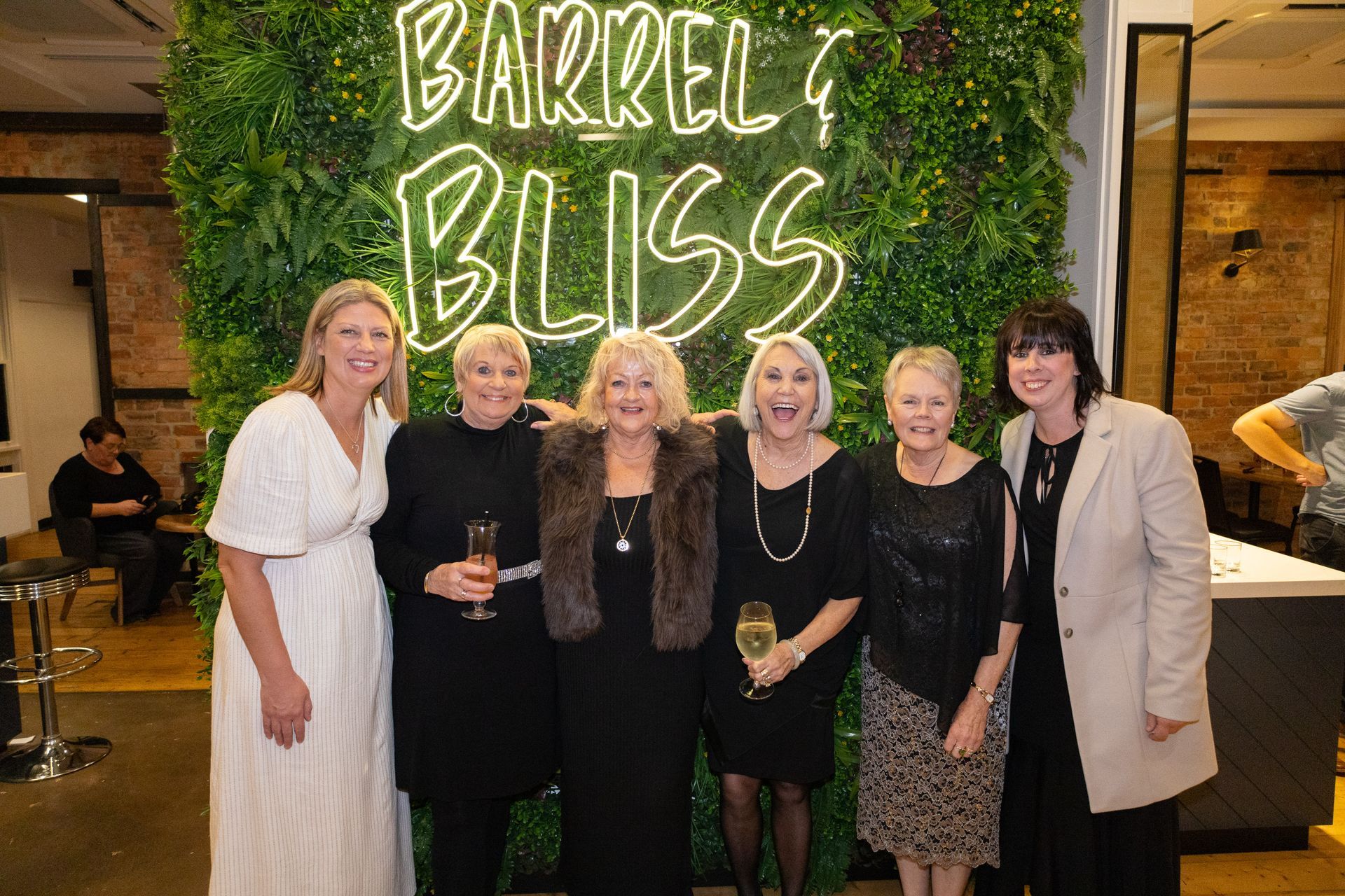 Six women smiling, posing in front of a neon sign that says 