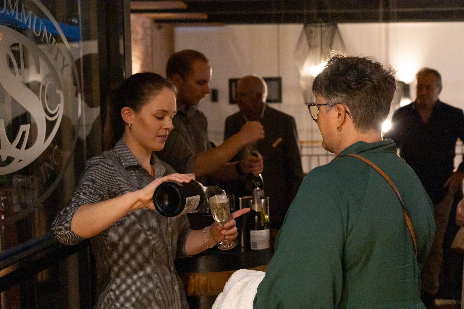 Woman in checkered shirt pours champagne for a guest at a restaurant, with others in the background.