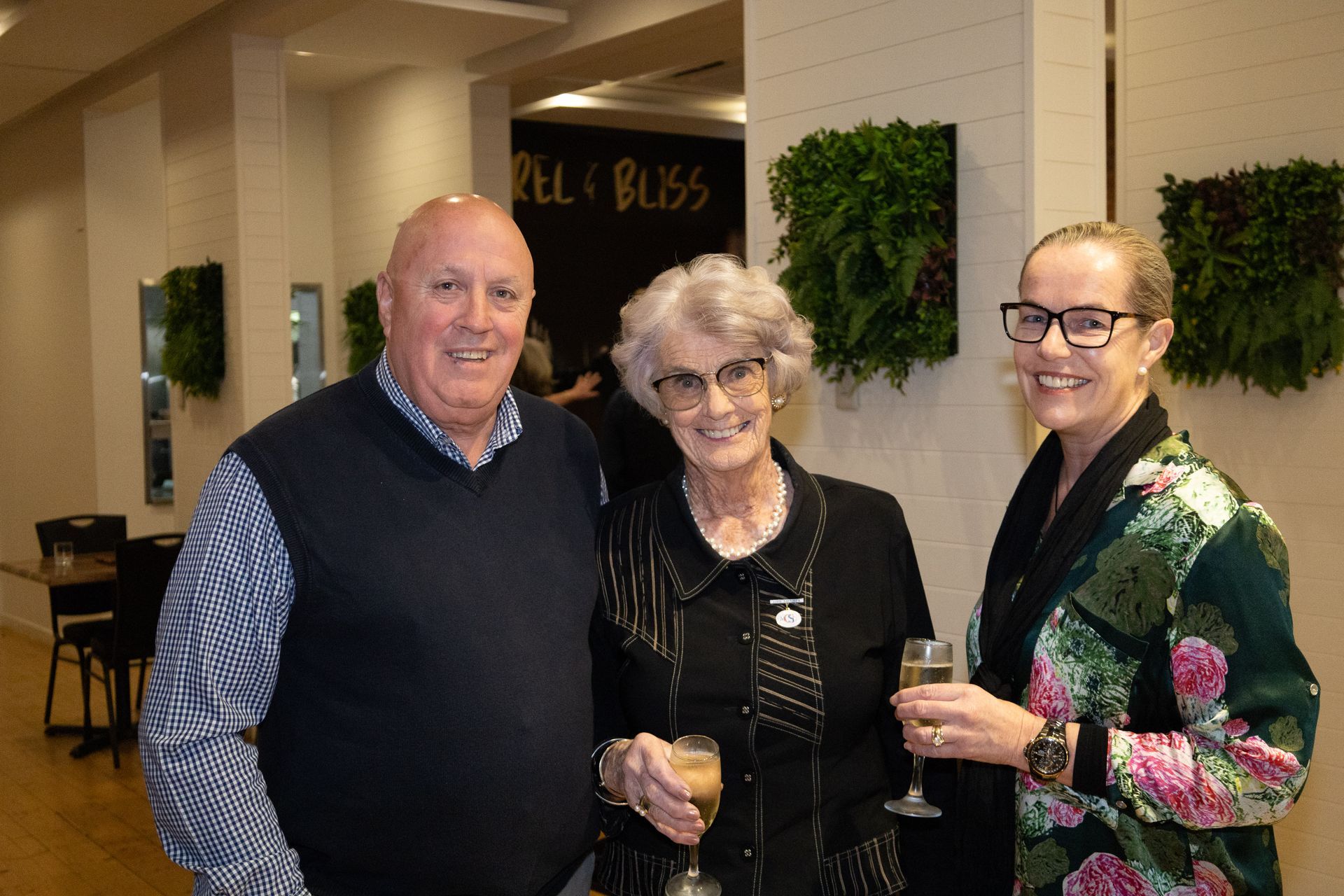 Three people smiling indoors at an event, posing near living plant wall decor.