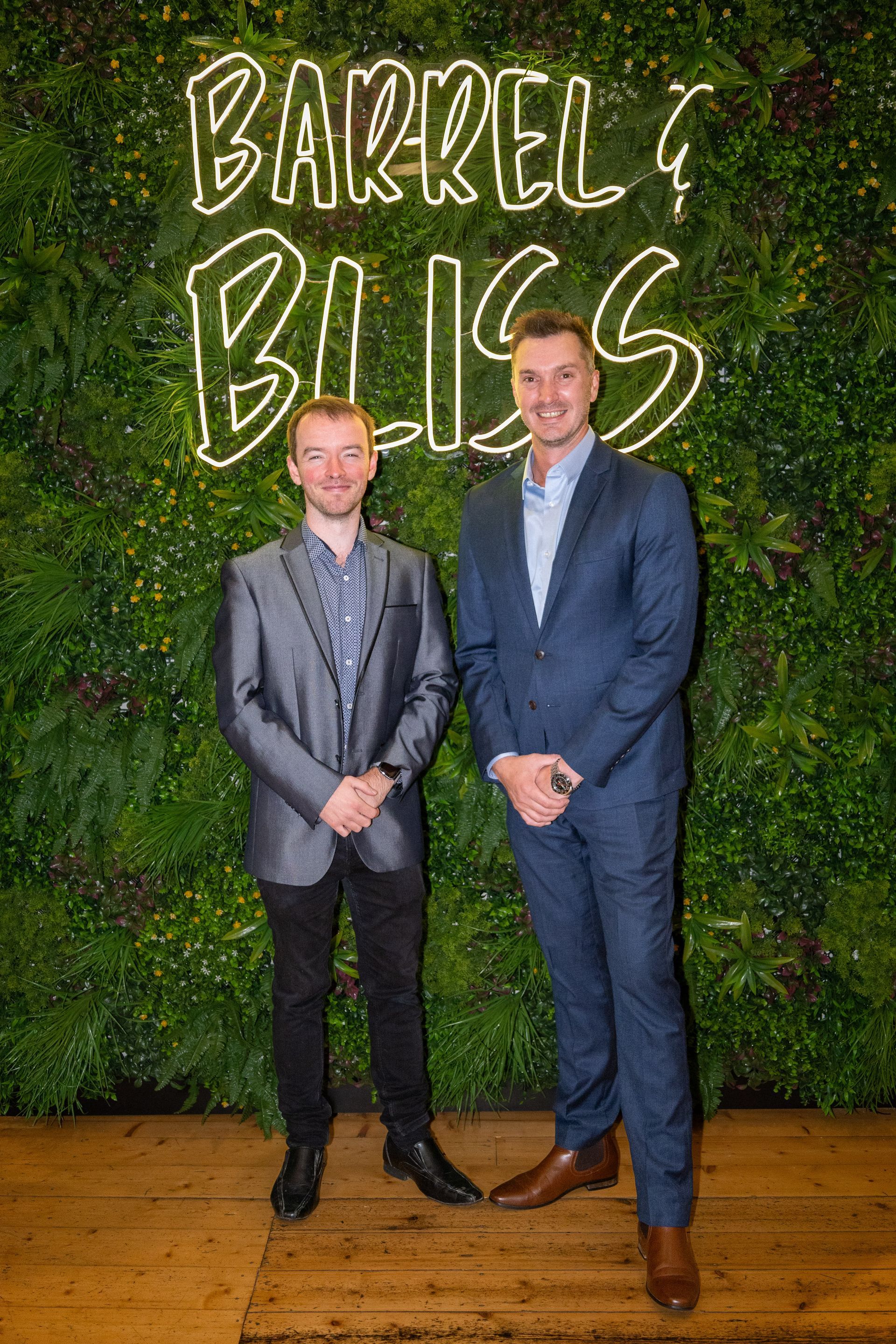 Two men in suits stand in front of a green wall with neon 