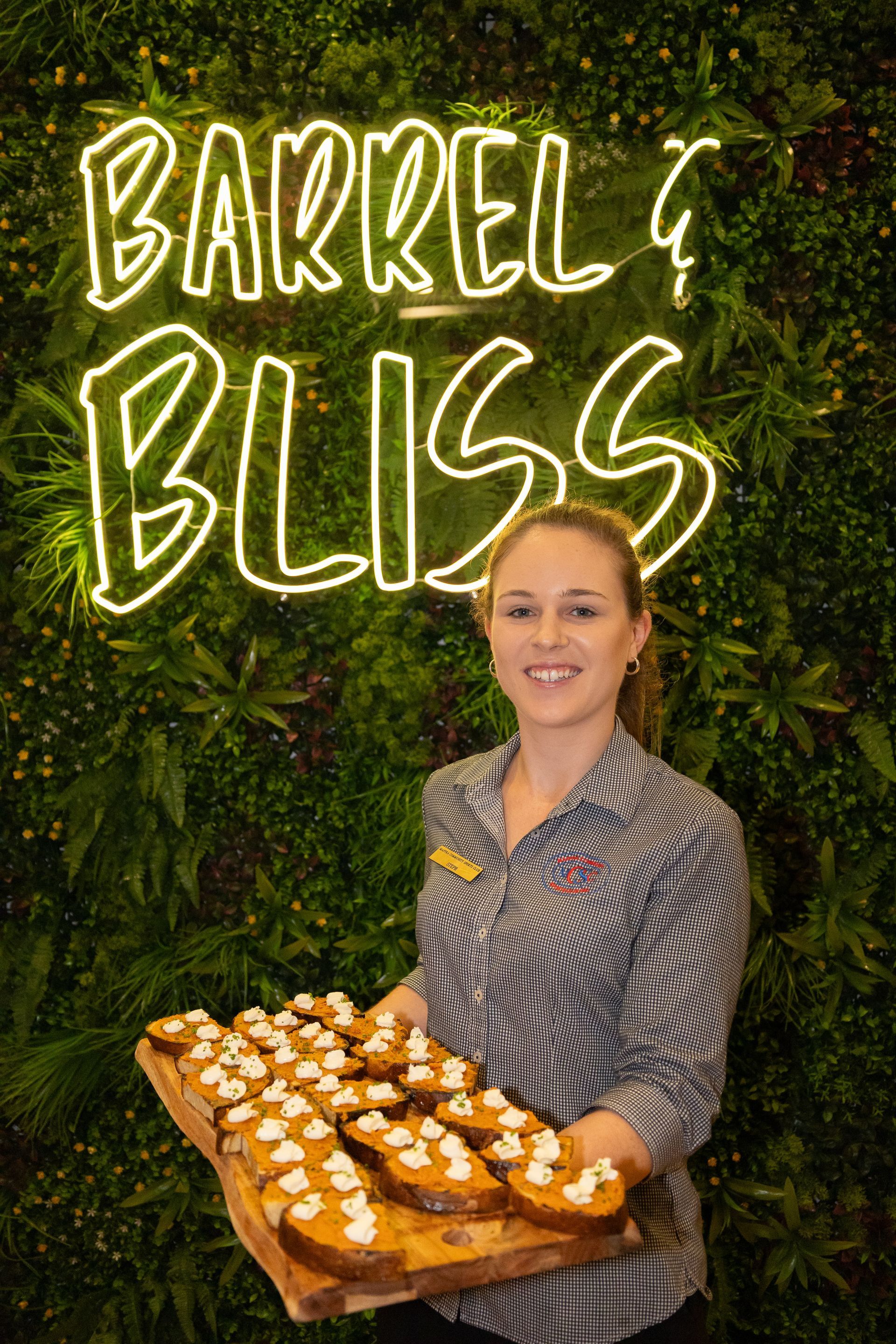Woman holding tray of food in front of a green wall with a neon sign that says “Barrel & Bliss”.