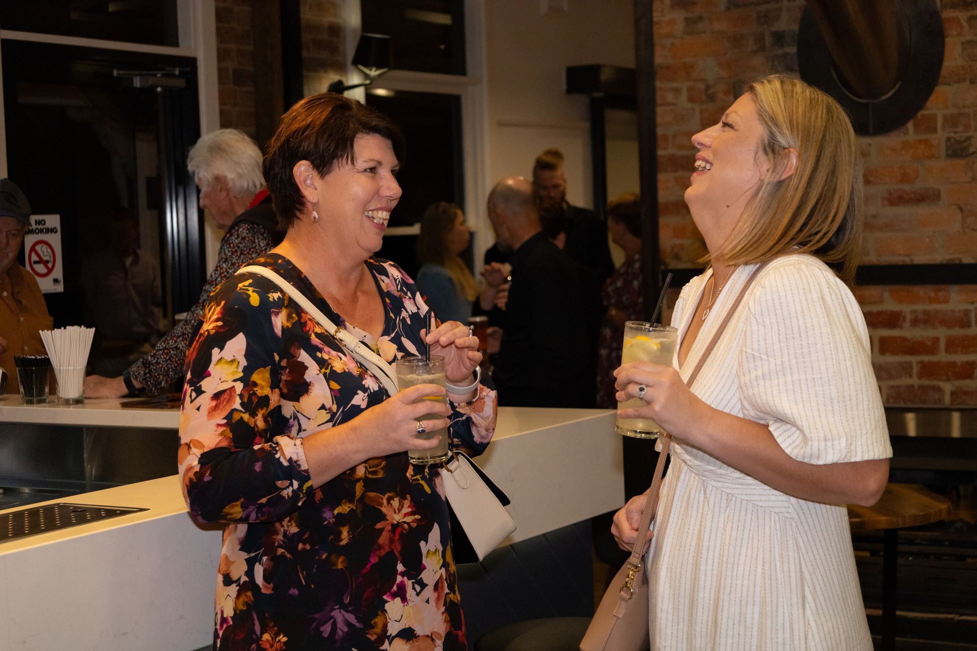 Two women laughing and holding drinks in a bar with a crowd in the background.
