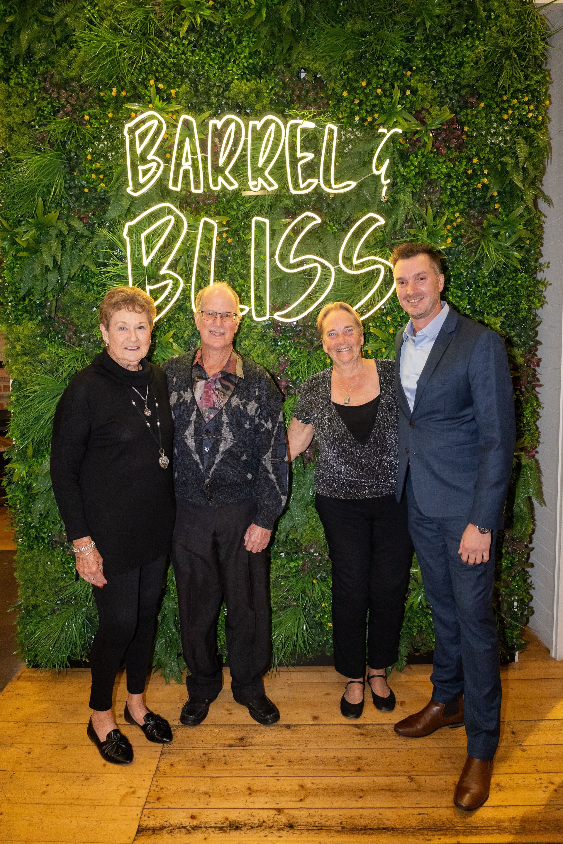 Four people pose in front of a green wall with a neon sign.