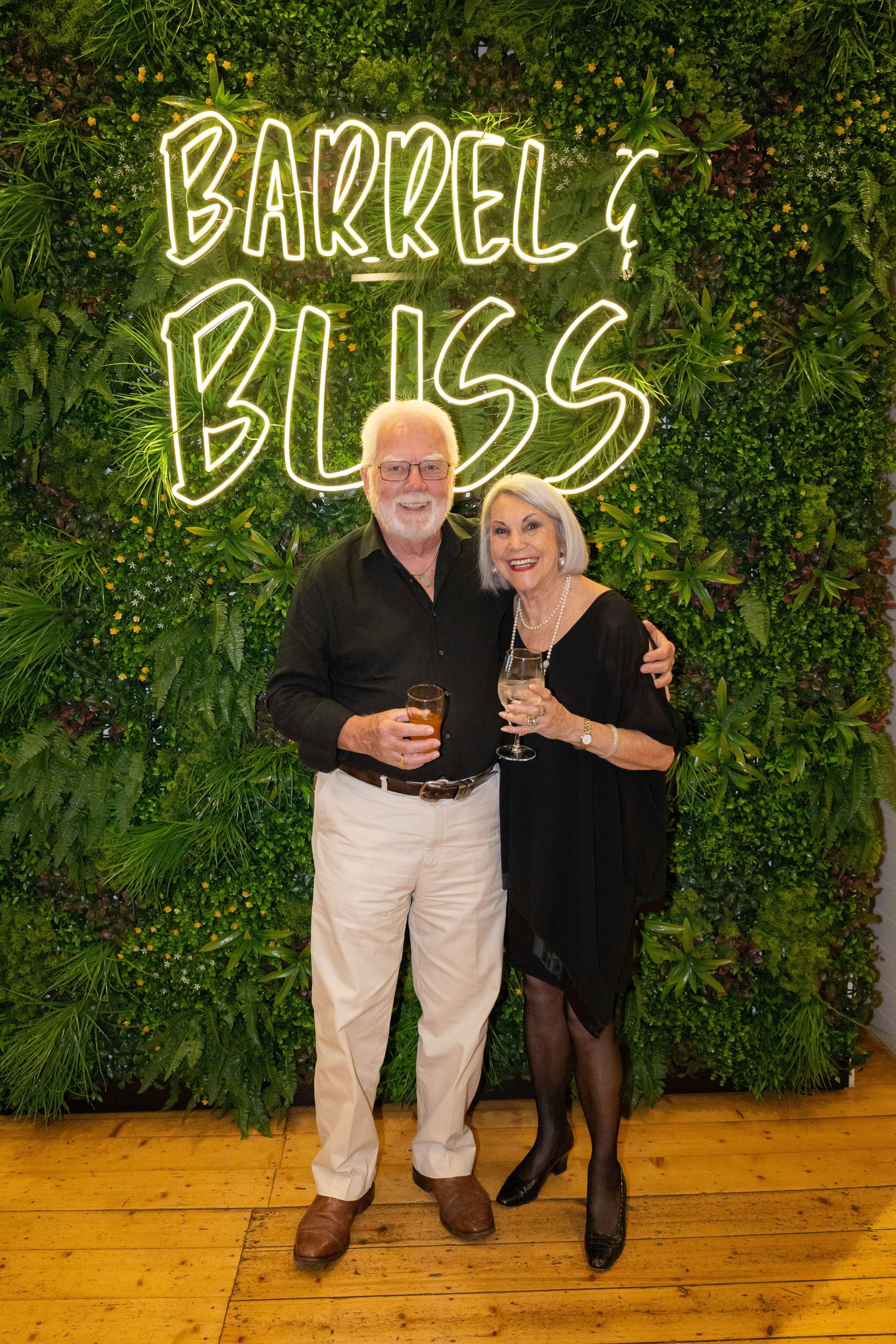 Couple smiling, holding drinks in front of a green wall with neon 