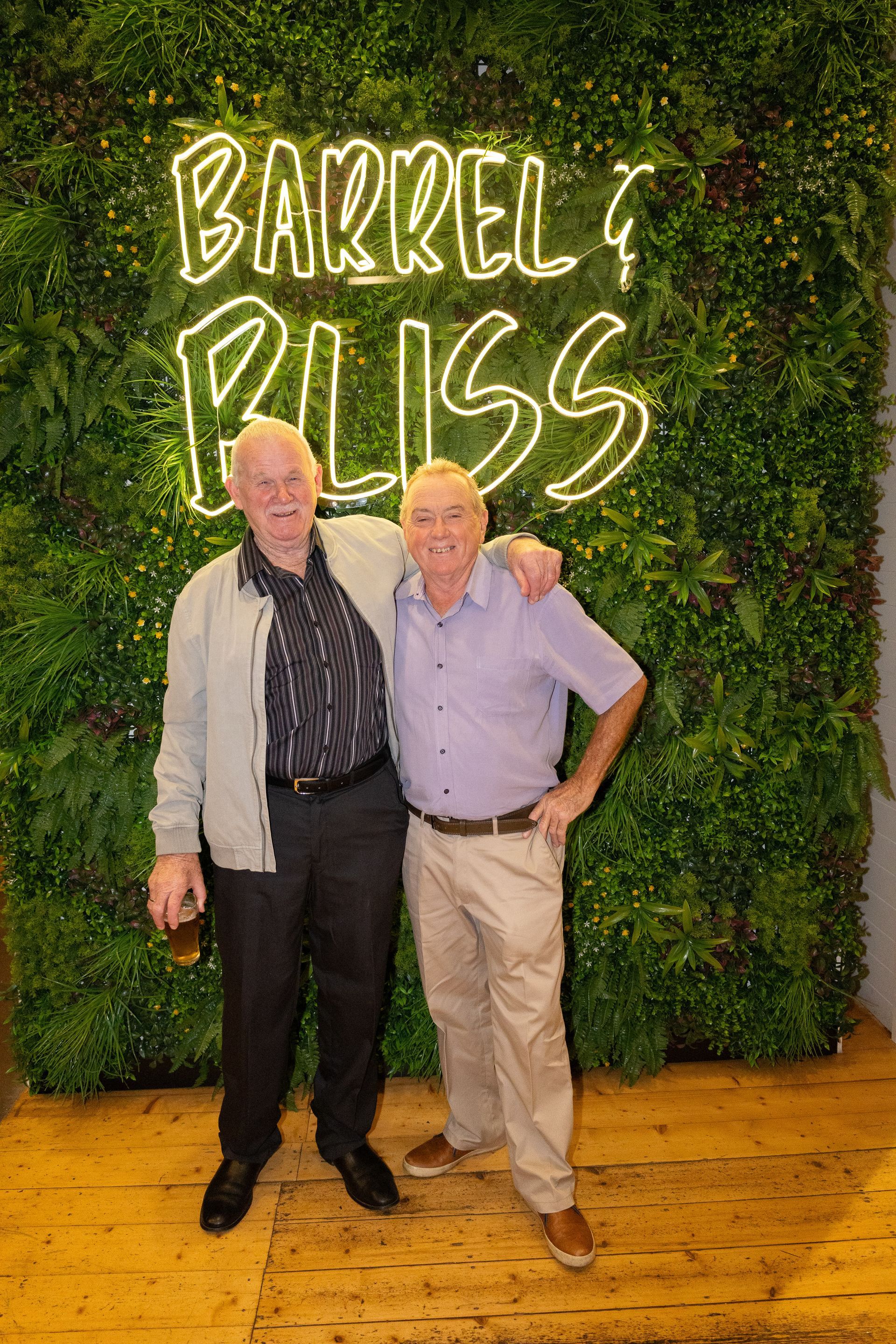 Two men pose in front of a neon sign that says 