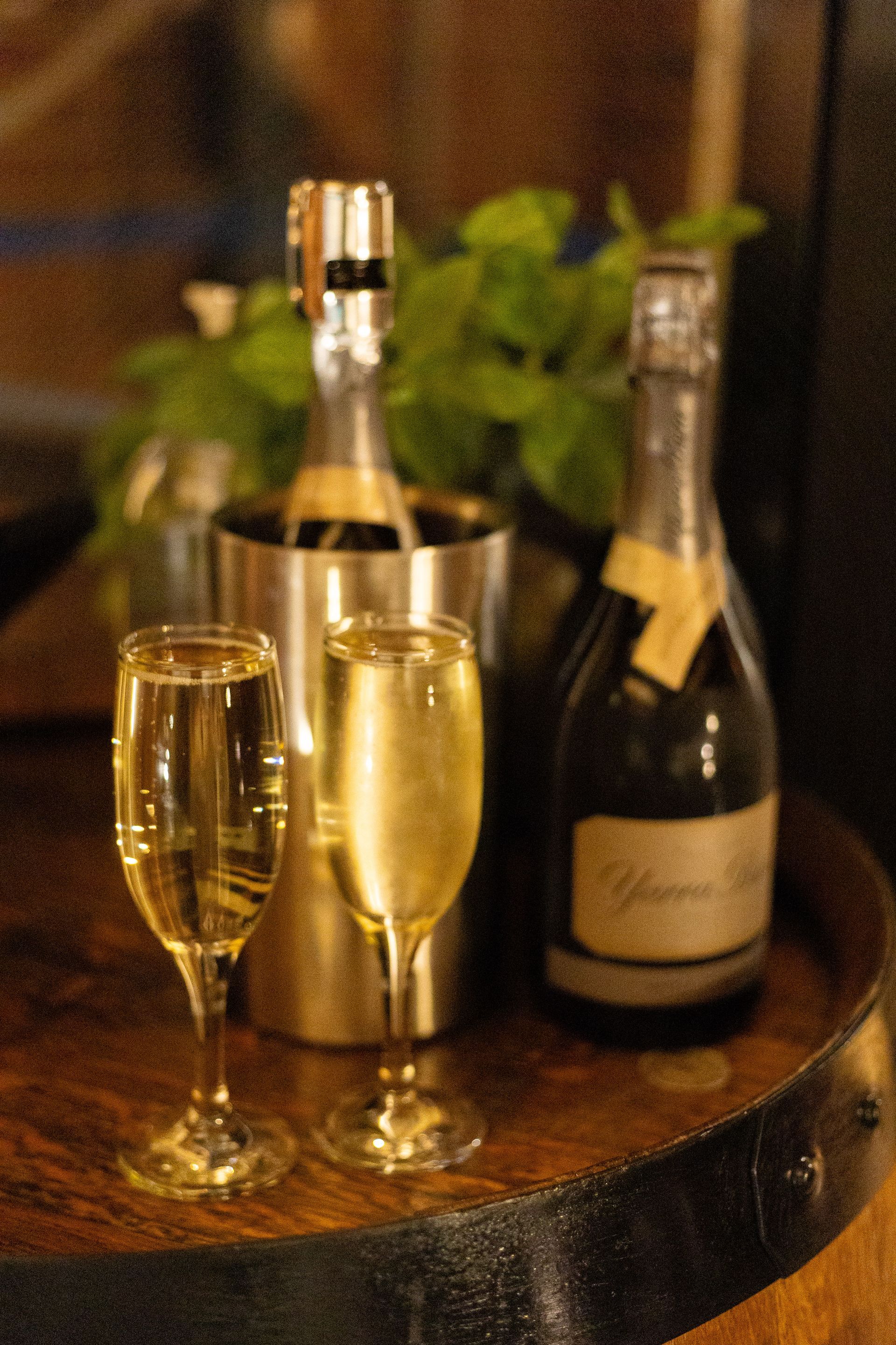 Two champagne flutes and two bottles of champagne on a wooden barrel; one bottle in an ice bucket.