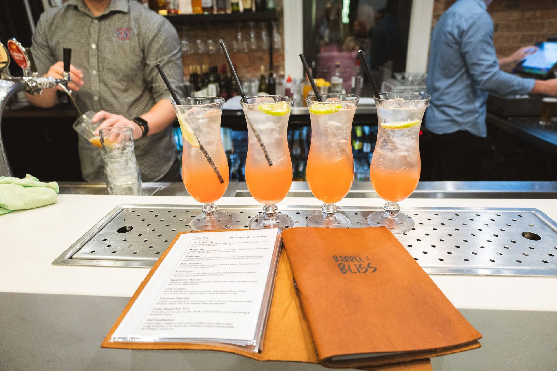 Bartender pouring a drink with four cocktails on the bar. Open menus are in front.