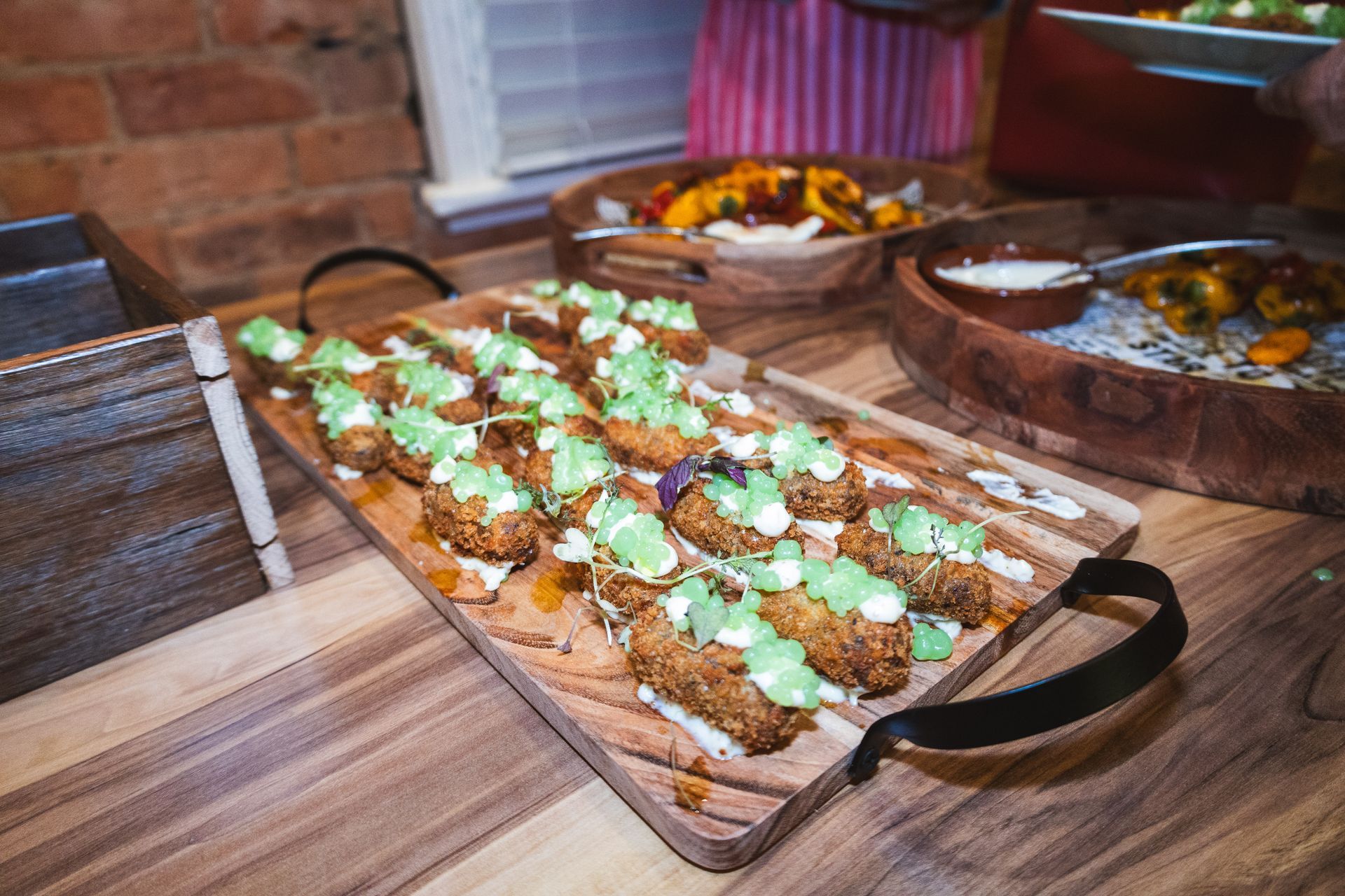 Wooden tray with appetizers topped with green sauce, on a wood table. Other food in background.