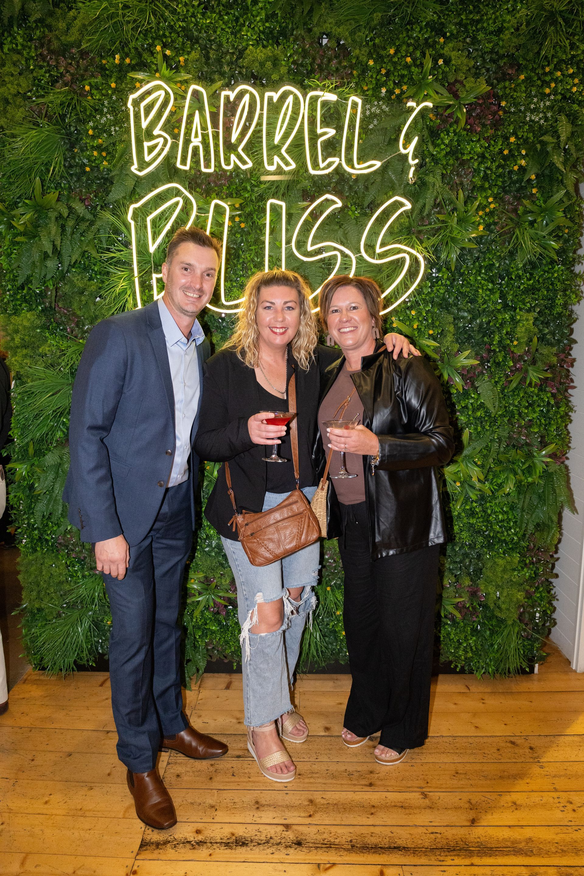 Three people smiling in front of a wall of plants and neon sign reading 