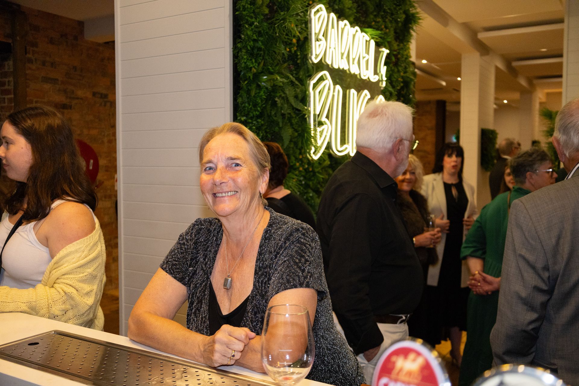 Woman at a bar smiles; Barrel & Burger sign in the background. Other people are in the room.